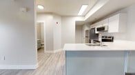 This single-floor unit offers a thoughtfully designed layout with a contemporary aesthetic defined by neutral tones, clean lines, and durable finishes throughout. The open-concept kitchen serves as a central feature, anchored by a peninsula with a white countertop and an integrated sink, complemented by white cabinetry, stainless steel appliances — including a refrigerator, electric range, and over-the-range microwave — and recessed panel lighting overhead. Light wood-style luxury vinyl plank flooring extends through the kitchen and living areas, lending warmth and continuity across the space.

The living area is generously proportioned, featuring neutral walls, large baseboards, and a sliding glass door fitted with vertical blinds that allows natural light to fill the room. Two interior doors lead to additional rooms, maintaining a logical and accessible flow throughout the unit.

The bedrooms are finished with plush neutral carpet and feature windows with horizontal blinds, providing a quiet, comfortable atmosphere. The bathroom includes a white vanity, a tub-shower combination with a curtain rod, and the same wood-style flooring found in the main living spaces. A secondary bathroom is also accessible from the main hallway. Overall, the space presents a clean, move-in-ready environment with consistent modern finishes.