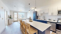 Located at 105 E Turner Mill Rd in Heber City, Utah, this single-floor residence combines modern design with functional living. The open-concept kitchen is a standout feature, showcasing bold navy-blue lower cabinetry paired with crisp white upper cabinets, a white quartz island with bar seating, a classic subway tile backsplash, and stainless steel appliances including a full-size refrigerator, range, and microwave. Pendant lighting and matte black hardware add refined detail throughout. The kitchen flows seamlessly into the living area, which features light wood-style flooring, large windows, and a glass-panel door that brings in generous natural light. The living room offers a comfortable, open layout well-suited for both relaxing and entertaining. The primary bedroom is a tranquil retreat with plush carpeting, a ceiling fan, and a window with exterior views. It is complemented by a spacious walk-in closet with wrap-around shelving and hanging rods. The en-suite primary bathroom features a dual-sink vanity with navy cabinetry, a large mirror, and wood-look flooring. A second bedroom provides a bright, serene space with its own large window. The secondary full bathroom continues the navy-blue vanity theme and includes a shower-tub combination with matte black fixtures and built-in shelving.