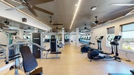 Located in Huntsville, Alabama, this well-appointed fitness center offers a generously sized, single-floor layout designed to support a full range of workout activities. The main exercise room is the centerpiece of the facility, featuring light hardwood-style flooring, crisp white walls, and a dark ceiling accented by sleek linear LED lighting. Large ceiling fans are distributed throughout, ensuring air circulation during intense workouts. Floor-to-ceiling mirrors line key wall sections, visually expanding the space and including a mounted large-screen display for classes or virtual training. Natural light flows in through a series of windows that frame views of the surrounding landscape, including lush greenery and distant hills. The cardio zone is equipped with treadmills, ellipticals, a rowing machine, and a stair climber, while the strength area houses a multi-station cable machine, leg press, and additional resistance equipment. A dedicated studio room offers space for cycling, functional training, and group exercise, stocked with spin bikes, kettlebells, medicine balls, stability balls, and foam rollers. A hallway connects the facility's zones, completing a thoughtfully organized and inviting athletic environment suited for comprehensive fitness routines.