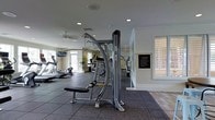 This single-level fitness center offers a thoughtfully designed workout space equipped with a comprehensive selection of exercise machines. The strength training area features a multi-functional cable machine with an adjustable bench, while the cardio section includes treadmills and additional aerobic equipment. Large shuttered windows allow natural light to filter into the space, contributing to a bright and welcoming atmosphere, while recessed lighting and ceiling fans ensure consistent illumination and air circulation throughout. The rubber-tile flooring provides both safety and durability underfoot, covering the main workout areas. A large mirror is positioned along one wall, enhancing the sense of depth and allowing users to monitor their form during exercise. A rules and guidelines board is mounted near the entrance, keeping users informed of facility policies. The neutral white walls and clean finishes give the space a polished, modern aesthetic. Overall, the fitness center balances practicality with comfort, providing an organized and well-maintained environment suitable for a range of workout routines.