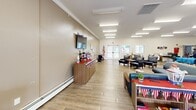 Located at 600 S Horner St in Lebanon, Illinois, this single-floor community space offers a generously sized, open-plan living and gathering area designed to accommodate a variety of social activities. The interior is finished with warm beige walls accented by crisp crown molding and light wood-tone flooring throughout, creating a clean and welcoming atmosphere.

The space is thoughtfully organized into distinct functional zones. A comfortable lounge area features navy blue upholstered armchairs and sofas arranged around small accent tables, ideal for relaxed conversation. A wall-mounted flat-screen television anchors one side of the room, complemented by a console unit below. A piano positioned along the back wall adds a touch of character and serves as a focal point for entertainment. Dining tables with chairs are arranged across another section of the room, accommodating group meals and gatherings.

A kitchenette or service counter area is visible toward the rear of the space, providing convenient support for events and activities. Multiple sets of exterior glass-panel doors line the perimeter walls, inviting ample natural light and offering views of the surrounding outdoor environment. Recessed and flush-mounted fluorescent ceiling fixtures provide consistent, bright illumination throughout. The open layout and accessible design make this a highly functional and inviting community gathering space.