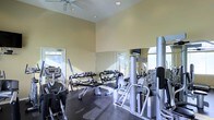 This fitness center is thoughtfully designed to support a comprehensive workout experience within a single-floor layout. The space features soaring ceilings that create an open, energizing atmosphere, complemented by large windows that flood the room with natural light. Warm, neutral-toned walls contribute to a clean and focused environment conducive to exercise.

The facility is well-stocked with a wide variety of equipment to accommodate diverse fitness needs. A full dumbbell rack lines one wall, providing a complete range of free weights for strength training. Multiple cable and weight machines, including a Vectra multi-station system, are positioned throughout the space for targeted muscle training. Cardio equipment, including an elliptical trainer, is also available for aerobic workouts.

Full-length mirrors span a significant portion of the walls, enhancing the sense of space while allowing users to monitor their form during exercise. The dark rubberized flooring provides both durability and safety underfoot. A ceiling fan ensures comfortable air circulation throughout workouts. Recessed lighting supplements the natural light, ensuring the space remains well-lit at all hours. Overall, this fitness center offers a functional and inviting environment for a complete training routine.