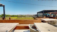This sophisticated rooftop amenity space offers a seamless blend of indoor comfort and outdoor living. The interior lounge area is designed with a warm, eclectic aesthetic, featuring a slatted wood ceiling, decorative lattice-patterned room dividers, and an array of plush seating arrangements anchored by woven pendant light fixtures in amber and geometric forms. Large floor-to-ceiling windows and sliding glass doors flood the interior with natural light while providing unobstructed views to the outdoor terrace beyond.

A dedicated bar area anchors one side of the interior, complete with a substantial granite-topped island, leather-upholstered bar stools with brass hardware, and a stone-clad accent wall housing flat-screen televisions. Glamorous cluster pendant lights add a refined touch overhead, while warm yellow ceiling tones lend a vibrant, energetic atmosphere to this entertainment-focused zone.

The outdoor terrace extends the usable space considerably, showcasing rich hardwood built-in bench seating arranged around concrete-top tables. A slatted pergola structure provides a shaded gathering area, while a sleek metal perimeter railing frames sweeping open views of the surrounding greenery and cityscape. Festoon string lights strung overhead create an inviting ambiance ideal for evening gatherings under the open sky.