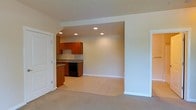 This single-floor space offers a clean, open-concept layout that brings together the living area and kitchen in a cohesive, functional design. The living area features soft, neutral-toned carpeting that creates a comfortable and inviting atmosphere, while the kitchen zone transitions to light ceramic tile flooring for a practical and polished look.

The kitchen is defined by warm medium-toned wood cabinetry that provides ample storage, complemented by dark granite-style countertops that add a touch of contrast and sophistication. A black appliance suite, including a range and dishwasher, contributes a modern, uniform aesthetic. The sink is positioned along the counter with easy access, and a subtle tile backsplash ties the kitchen elements together neatly.

Recessed lighting is installed throughout both the living and kitchen areas, providing even, well-distributed illumination across the entire space. The transition between the two areas is marked by a subtle architectural soffit, giving the open floor plan a sense of structure without sacrificing flow.

White panel doors and clean white baseboards frame the space, reinforcing the bright and well-maintained character of the interior. The overall ambiance is understated and move-in ready, with a neutral palette that offers flexibility for a variety of furnishing styles.