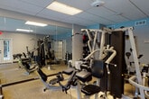 This fitness center is a comprehensively outfitted exercise facility designed to support a full range of workout routines. The space is organized into distinct functional zones, maximizing the available floor area for both cardio and strength training activities. A drop ceiling with recessed panel lighting and ceiling fans ensures consistent, comfortable illumination throughout.

The cardio zone features a row of treadmills and an elliptical machine positioned along a windowed wall. Large multi-pane windows overlook a lush outdoor area with a visible pool, filling this section with natural light and offering a pleasant view during workouts. A wall-mounted flat-screen television is positioned nearby for entertainment.

The strength training area is equipped with multiple cable and resistance machines, including a multi-function cable system, leg press, and additional weight stations. A dumbbell rack and free weights are also available. Full-length mirrors line the walls, visually expanding the space and allowing users to monitor their form. The durable speckled flooring provides cushioning and traction throughout.

French doors open directly to an outdoor patio, offering convenient access and ventilation. A separate entry vestibule with tile flooring provides a clean transition point into the main exercise area. A spin bike and step platforms round out the facility's versatile equipment offerings.