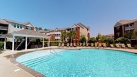 Located in Spanish Fort, Alabama, this residential community presents an inviting outdoor environment centered around a sparkling resort-style swimming pool. The pool area is generously sized, featuring clear blue water and a classic curved design that serves as the focal point of the outdoor amenity space. Surrounding the pool is a spacious sundeck lined with lounge chairs, ideal for relaxing under the region's abundant sunshine. A pergola-covered structure on the side of the pool deck provides a shaded retreat, adding both function and visual interest to the outdoor space. The community buildings feature a traditional architectural style characterized by brick and neutral-toned siding, gabled rooflines, and private balconies that overlook the grounds. Lush landscaping, including mature trees and manicured hedgerows, frames the property and contributes to a sense of privacy and greenery throughout. The overall ambiance is warm and resort-inspired, blending classic Southern residential architecture with thoughtfully designed outdoor amenities. Under clear blue skies, the community exudes a bright, open atmosphere well-suited to an active and relaxed lifestyle in the Spanish Fort area.