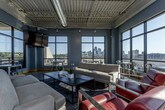 This single-floor space showcases a striking industrial aesthetic defined by exposed concrete floors, textured spray-foam ceilings, visible ductwork, and open structural elements that lend an authentic urban character throughout. The expansive main lounge area is bathed in abundant natural light, courtesy of large black-framed floor-to-ceiling windows that frame impressive panoramic city skyline views on multiple sides. The lounge is thoughtfully furnished with a pair of neutral sofas, bold red accent chairs, and a sleek rectangular coffee table anchored by a dark area rug, creating a comfortable yet contemporary gathering space. A wall-mounted flat-screen TV and additional high-top tables and bistro seating along the windowed perimeter offer versatile options for both relaxation and informal meetings. Adjacent to the lounge, the full kitchen is outfitted with dark cabinetry, light stone countertops, stainless steel appliances including a full-size refrigerator and built-in microwave, and a spacious white island with bar stool seating. The open floor plan allows the lounge and kitchen areas to flow seamlessly into one another, reinforcing the space's communal, collaborative atmosphere. The overall ambiance is modern and energetic, perfectly balancing industrial raw materials with polished, functional design.