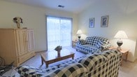 This single-floor space centers around a comfortable living room that draws immediate attention with its large sliding glass door, dressed in vertical blinds that diffuse abundant natural light throughout the room. The soft, neutral-toned carpet underfoot adds warmth and comfort to the space.

The living room is furnished with a coordinated set of black-and-white checkered sofas arranged around a solid wood coffee table, which serves as the focal point of the seating area. A decorative floral arrangement on the coffee table adds a touch of color to the classic, traditional aesthetic. The furniture arrangement encourages conversation and relaxed gathering.

Framed botanical artwork adorns the walls, complementing the traditional decor, while a table lamp with a classic shade provides warm supplemental lighting alongside the natural light from the sliding doors. A freestanding wooden armoire positioned near the entrance adds practical storage and visual character.

The overall ambiance of the space is calm and traditional, with a cohesive design scheme that prioritizes comfort and functionality. The sliding glass door hints at outdoor access, adding a sense of openness to the interior.