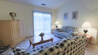 This single-floor space centers around a comfortable living room that draws immediate attention with its large sliding glass door, dressed in vertical blinds that diffuse abundant natural light throughout the room. The soft, neutral-toned carpet underfoot adds warmth and comfort to the space.

The living room is furnished with a coordinated set of black-and-white checkered sofas arranged around a solid wood coffee table, which serves as the focal point of the seating area. A decorative floral arrangement on the coffee table adds a touch of color to the classic, traditional aesthetic. The furniture arrangement encourages conversation and relaxed gathering.

Framed botanical artwork adorns the walls, complementing the traditional decor, while a table lamp with a classic shade provides warm supplemental lighting alongside the natural light from the sliding doors. A freestanding wooden armoire positioned near the entrance adds practical storage and visual character.

The overall ambiance of the space is calm and traditional, with a cohesive design scheme that prioritizes comfort and functionality. The sliding glass door hints at outdoor access, adding a sense of openness to the interior.