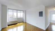 This single-floor residence offers a clean, minimalist aesthetic defined by crisp white walls and polished parquet wood flooring in a classic herringbone pattern that adds warmth and timeless character to the interior. The main living area is generously proportioned, benefiting from an abundance of natural light streaming through large, floor-to-ceiling windows. These expansive windows frame sweeping elevated views of the surrounding cityscape, suggesting a high-floor position within a larger urban building. A baseboard heating unit is positioned beneath the windows, ensuring year-round comfort. The bright, open feel of the space is enhanced by the seamless white ceiling and well-maintained walls. An electrical panel box is neatly mounted on one wall, representing a practical built-in feature of the unit. The overall ambiance is airy and uncluttered, providing a versatile blank canvas well-suited for a variety of interior design styles. The combination of natural light, city views, and classic architectural flooring details make this a compelling urban living space with both functional and aesthetic appeal.