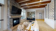 Located at 4027 Watercraft Ferry Avenue in Wilmington, NC, this thoughtfully designed single-floor space blends modern aesthetics with warm, inviting details throughout. The main lounge area draws immediate attention with its striking stone-clad fireplace, wall-mounted TV, and richly toned wood beam ceiling that adds architectural depth and warmth overhead. Large floor-to-ceiling windows and a glass-panel entry door flood the interior with natural light while offering views of the surrounding outdoor greenery. Herringbone-patterned hardwood flooring flows throughout, grounding the space with a refined, cohesive look. A decorative laser-cut lattice screen partition elegantly defines zones within the open floor plan without compromising the sense of spaciousness. A cozy lounge area features plush seating and textured poufs, while a built-in shelving unit with teal-backed display niches and a bistro-style table creates an intimate nook for reading or casual conversation. The kitchen area is a standout, showcasing vibrant aqua-blue lower cabinetry paired with white quartz countertops and a striking geometric mosaic tile backsplash that adds visual texture. Open shelving above keeps the aesthetic light and airy. Together, these elements create a dynamic, design-forward space that is both functional and visually engaging.