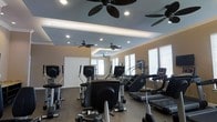 This dedicated fitness space is thoughtfully designed to support a full cardio workout experience. The room's most striking architectural feature is its tiered tray ceiling, finished in a cool blue-gray tone and accented with recessed lighting that provides bright, even illumination throughout. Multiple ceiling fans are strategically positioned across the ceiling tiers, ensuring consistent airflow and comfort during exercise sessions.

The walls are finished in a warm neutral tone, complemented by white crown molding that adds a polished, refined touch. Several windows fitted with white horizontal blinds allow natural light to filter into the space while maintaining privacy and light control.

The fitness area is generously sized, accommodating a diverse collection of cardio equipment including elliptical trainers, stair climbers, and treadmills, all arranged to allow comfortable circulation between machines. The flooring appears to be a durable, low-pile carpet suitable for a high-traffic exercise environment. A white paneled door provides access to an adjoining room or corridor. Overall, the space balances functionality with a clean, well-maintained aesthetic.