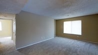 This single-story space presents a straightforward, functional layout centered around a spacious open room. The walls are finished in a warm, neutral tan tone that complements the wall-to-wall carpeting underfoot, creating a cohesive and understated aesthetic throughout. The flooring is plush carpet in a light gray-beige shade, offering comfort and a soft visual continuity across the space.

The ceiling features a textured popcorn finish, a common characteristic of its construction era, and includes a ceiling vent for climate control. White baseboards run along the perimeter of the room, providing a clean contrast against the tan walls and carpet.

A single window on the far wall allows natural light to filter into the room, fitted with horizontal blinds that offer privacy and light control. The room's generous open floor plan provides flexibility for a variety of furniture arrangements and uses.

Electrical outlets are conveniently positioned along the walls, adding to the room's practical utility. Overall, the space maintains a clean, move-in-ready condition with a calm, neutral palette that serves as a blank canvas for personalization.