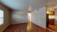 This single-floor unit is characterized by consistent warm-toned hardwood-style flooring that runs throughout every room, lending a cohesive and inviting feel to the entire space. The layout, as seen in the floor plan, includes a generously sized living area, a separate kitchen, a full bathroom, and multiple additional rooms suitable for use as bedrooms or flexible living spaces.

The living area is open and well-proportioned, with neutral-painted walls, standard ceilings, and baseboard heating units along the walls. A front entry door and a window with blinds allow natural light to fill the space, providing views of the surrounding exterior.

The kitchen features warm wood cabinetry with ample upper and lower storage, granite-style countertops, a stainless steel single-basin sink, and a black range. A white subway tile backsplash adds a clean, functional accent behind the cooking and prep areas.

The bathroom is bright and neatly appointed, with a full tub-shower combination, a white vanity with under-cabinet storage, a wall mirror, and matching hardwood-style flooring.

The additional rooms each include windows with blinds that bring in natural light, neutral walls, and the same continuous flooring, offering versatile and functional spaces throughout the unit.