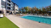 Located at 1440 Yellow Desert Way in Wendell, NC, this outdoor amenity space delivers a resort-style experience designed for relaxation and recreation. At the heart of the area is a generously sized swimming pool with crystal-clear water, complete with in-pool sun shelves and water features that add a dynamic, inviting atmosphere. The pool deck is finished with elegant paver brickwork, offering ample space for lounging alongside a collection of chaise chairs and shaded umbrella stations. Adjacent to the main pool, a separate shallow wading area provides a secondary space for leisure, also surrounded by lounge seating. A dedicated turf lawn area offers a lush, green alternative for sunbathing, lined with additional chaise loungers and framed by black metal fencing. A warm wood-toned pergola provides a shaded gathering spot, ideal for outdoor dining or socializing. Thoughtfully landscaped planting beds with ornamental grasses and shrubs add texture and greenery throughout the space. The surrounding multi-story residential building, clad in a combination of brick, dark siding, and large windows, encloses the area and creates a sense of privacy. Mature trees along the perimeter provide natural backdrops and partial shade, enhancing the open-air ambiance under the bright North Carolina sky.