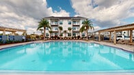 The pool area is a thoughtfully designed outdoor amenity space centered around a generously sized swimming pool with crystal-clear turquoise water. The pool's smooth, curvilinear edges are complemented by a surrounding deck, while tall palm trees frame the space and reinforce the tropical ambiance. A multi-story residential building with white exterior siding and private balconies serves as an elegant backdrop.

Adjacent to the pool, a covered pavilion provides shaded outdoor living space finished with a durable, multi-toned stone paver floor. The pavilion is supported by substantial squared columns and features multiple distinct seating zones. One area offers dark wood dining tables and chairs suited for casual meals, while another section hosts a plush, neutral-toned sectional sofa arrangement ideal for lounging. The covered structure is enclosed partially by a black metal fence, offering both aesthetic definition and safety.

The entry to the pavilion area is marked by a glass-and-dark-metal door flanked by large sliding windows, with white shiplap siding adding a coastal architectural character. Vibrant potted plants with colorful foliage flank the entrance, providing welcoming, decorative greenery. The overall atmosphere is polished, relaxed, and oriented toward outdoor leisure and social gathering.