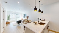 This thoughtfully designed one-bedroom, one-bathroom unit in Aurora, Colorado offers a clean, contemporary aesthetic with high-quality finishes throughout. The open-concept kitchen and living area serves as the heart of the home, anchored by a large central island topped with elegant marble-look quartz countertops and seating for multiple guests. A trio of striking matte black pendant lights with gold interiors hangs above the island, adding a bold design accent. The kitchen features flat-front cabinetry extending to the ceiling, stainless steel appliances, a herringbone tile backsplash, and a matte black faucet. Light hardwood flooring flows seamlessly through the main living area. The living room is bright and airy, benefiting from oversized windows that invite generous natural light and outdoor views. The bedroom offers plush carpeting, a ceiling fan, a dedicated closet, and its own window overlooking the surroundings. The full bathroom is well-appointed with a soaking tub and shower combination, a floating wood vanity, a large framed mirror, and black fixture accents. The overall ambiance is modern, minimal, and inviting — ideal for comfortable urban living.