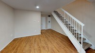 This two-story townhome offers a practical and comfortable layout across two levels, finished with warm hardwood-style flooring that runs consistently throughout. The lower level features an open living area with recessed lighting, neutral beige walls, and a staircase with a clean white railing leading to the upper floor. Two white interior doors on the ground floor provide access to additional spaces, including what appears to be a utility or storage area. A sliding glass door on this level opens to an outdoor area, allowing natural light to fill the space.

The upper level serves as the primary living and sleeping area, centered around an open room with a ceiling fan and a large picture window that frames outdoor views and draws in ample natural light. The white stair railing continues on this floor, maintaining a cohesive aesthetic. A frosted-glass exterior door and additional interior doors provide access to the bathroom and other areas.

The bathroom features a full tub-shower combination with a white surround, a toilet, and an over-the-toilet storage cabinet with paneled doors. Granite-style countertop detail is visible, adding a refined touch. Neutral tones and clean finishes give the entire townhome a bright, move-in-ready feel.