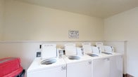 Located at 360 Broad Meadows Blvd in Columbus, Ohio, this single-floor shared laundry facility is designed to serve residents with convenience and practicality in mind. The main laundry area features a row of top-loading washing machines aligned along one wall, complemented by stacked dryer units positioned nearby, offering ample capacity for multiple users at once. A laundry cart is available for residents' use, and a folding table provides a dedicated surface for sorting and organizing clothes. Wood-look flooring runs throughout the space, lending a clean and durable finish underfoot, while textured ceilings and neutral-toned walls create a bright, utilitarian atmosphere. Fluorescent overhead lighting ensures the room is well-lit at all hours. Adjacent to the laundry area is a waiting space furnished with orange molded chairs and a long communal table, offering residents a comfortable place to wait. A cork bulletin board is mounted on the wall, serving as a community information hub. A soda vending machine rounds out the amenities. Posted signage indicates that laundry hours run from 8 AM to 10 PM, for residents only. A window in the laundry room allows for natural light, adding to the functional and welcoming character of the space.