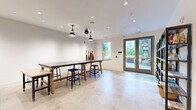 Located at 111 E Broad St in Falls Church, Virginia, this single-floor studio space is thoughtfully designed for creative and hands-on activities. The open layout centers around a large, dark-topped worktable with wooden trestle legs, accompanied by blue metal stools and a mobile utility table, offering ample workspace for a variety of projects.

Polished concrete floors run throughout, lending the space an industrial yet clean aesthetic that is both durable and easy to maintain. Overhead, a combination of recessed can lights and distinctive black industrial pendant fixtures — including a pulley-style lamp — provide layered, functional illumination suited to detailed work.

Natural light fills the room through multiple windows and a set of glass double doors that open directly onto an outdoor patio, offering pleasant views of the surrounding greenery. This seamless indoor-outdoor connection enhances the overall sense of openness.

Along one wall, a built-in kitchenette with cabinetry and a sink provides practical utility for cleaning up after projects. On the opposite side, a dedicated hallway area features floor-to-ceiling open metal shelving units stocked with art supplies, books, and tools, keeping materials organized and accessible.

Aprons hanging on wall hooks and the curated supply shelves reinforce this space's identity as a well-equipped, community-oriented creative studio in the heart of Falls Church.