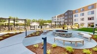 Located at 80 Batterson Park Rd in Farmington, Connecticut, this thoughtfully designed outdoor courtyard offers a well-appointed residential amenity space surrounded by a modern multi-story building. The grounds are meticulously landscaped, featuring expansive stretches of lush green lawn that serve as the central focal point of the courtyard. A stone fire pit with curved bench seating provides a welcoming gathering spot, while winding paved pathways connect the various zones throughout the space. Rattan egg chairs and wooden lounge chairs are strategically placed throughout, offering relaxed seating options amid the greenery. Raised garden beds and carefully curated plantings, including ornamental shrubs, young trees, and ground cover, add texture and visual interest to the landscape. A pergola and covered lounge area provide shaded retreats, complemented by market umbrellas nearby. The space also features access to a fenced outdoor pool area. The building exterior showcases a contemporary design with board-and-batten siding, large reflective windows, and mixed material facades in neutral tones. Architectural lighting elements are positioned throughout the courtyard, ensuring the space is equally functional and inviting after dark.