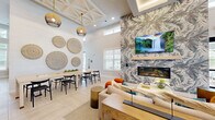 Located at 3100 Ascend Loop in Leland, NC, this single-story community clubhouse offers a thoughtfully designed gathering space that blends coastal-inspired aesthetics with modern functionality. The open-plan social lounge serves as the heart of the facility, featuring soaring ceilings with decorative coffered details, recessed lighting, and light-toned large-format tile flooring laid in a herringbone pattern. A dramatic accent wall clad in botanical-patterned wallcovering anchors a sleek linear fireplace beneath a wall-mounted television, creating a warm and inviting focal point. Comfortable seating arrangements, rattan accent chairs, and gold-toned furnishings contribute to the refined coastal ambiance throughout. The game room provides a lively recreational atmosphere, complete with a pool table, shuffleboard, wall-mounted displays, and striped banquette seating set within alcoves. A dedicated conference room features black-framed glass partition doors, a long natural wood table, and designer pendant lighting, making it well-suited for meetings or private gatherings. A fully equipped kitchen/bar area supports entertaining needs, while hallways connect the various functional zones seamlessly. The expansive covered outdoor patio extends the living space outdoors, offering a stacked-stone fireplace feature, ceiling fans, outdoor seating, and open views of the surrounding community and landscaping.