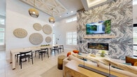 Located at 3100 Ascend Loop in Leland, NC, this single-story community clubhouse offers a thoughtfully designed gathering space that blends coastal-inspired aesthetics with modern functionality. The open-plan social lounge serves as the heart of the facility, featuring soaring ceilings with decorative coffered details, recessed lighting, and light-toned large-format tile flooring laid in a herringbone pattern. A dramatic accent wall clad in botanical-patterned wallcovering anchors a sleek linear fireplace beneath a wall-mounted television, creating a warm and inviting focal point. Comfortable seating arrangements, rattan accent chairs, and gold-toned furnishings contribute to the refined coastal ambiance throughout. The game room provides a lively recreational atmosphere, complete with a pool table, shuffleboard, wall-mounted displays, and striped banquette seating set within alcoves. A dedicated conference room features black-framed glass partition doors, a long natural wood table, and designer pendant lighting, making it well-suited for meetings or private gatherings. A fully equipped kitchen/bar area supports entertaining needs, while hallways connect the various functional zones seamlessly. The expansive covered outdoor patio extends the living space outdoors, offering a stacked-stone fireplace feature, ceiling fans, outdoor seating, and open views of the surrounding community and landscaping.