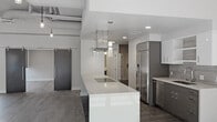 Located in the heart of Downtown Los Angeles, this modern single-floor residence at 225 S Olive St offers a thoughtfully designed interior with contemporary finishes throughout. The open-concept kitchen serves as a central hub, featuring a large waterfall-edge island with an induction cooktop, a stainless steel refrigerator, dark cabinetry, white upper cabinets with open shelving, a tile backsplash, and a statement stainless steel range hood. Pendant lighting and recessed fixtures illuminate the space, which transitions seamlessly into the living areas via wood-look flooring. A distinctive sliding barn door adds a modern industrial touch. The living and dining areas benefit from large floor-to-ceiling windows and sliding glass doors that open to a balcony, flooding the rooms with natural light and offering urban skyline views. Light wood flooring and warm solar shades complement the bright, airy atmosphere. Bedrooms are finished with plush carpeting, ceiling fans, and expansive windows with city views. The bathrooms are elegantly appointed with large-format tile, glass-enclosed soaking tubs, mosaic accent tile, white quartz countertops, and gray cabinetry. A powder room with clean, minimal finishes rounds out this well-appointed urban residence.