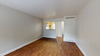 This two-floor space offers a practical and functional layout with a clean, neutral aesthetic throughout. The main living area is generously sized, finished with rich, dark hardwood-style flooring that extends across the open floor plan. Neutral gray walls and white trim create a bright, understated backdrop, while floor-mounted vents indicate a central HVAC system. A pass-through opening connects the living area to the kitchen, which features warm wood cabinetry, granite-look countertops, a double stainless steel sink, and a window that allows natural light and outdoor views into the space. A dishwasher is also present in the kitchen.

A separate entry area includes a front door with an adjacent window, bringing additional natural light into the home. The bedroom is carpeted and offers a ceiling fan with lighting, along with a window framing views of mature trees. The full bathroom is equipped with a tub-shower combo, toilet, wood vanity, granite-look countertop, and a large mirror with overhead lighting.

The lower level consists of an unfinished basement with a painted concrete floor and exposed ceiling joists, housing the water heater and furnace — providing functional utility space with potential for additional use.