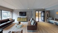 This community room is a thoughtfully designed, open-plan gathering space that seamlessly blends multiple functional zones into one cohesive environment. The lounge area serves as a focal point, anchored by a dramatic floor-to-ceiling stone tile accent wall on which a large flat-screen television is mounted. A gold-framed media console sits beneath the TV, complementing the rich brown leather sofas and accent chairs arranged around a marble-topped, gold-base coffee table atop a textured area rug.

Adjacent to the lounge, a substantial reclaimed wood dining table with upholstered chairs accommodates group gatherings or collaborative work sessions. A smaller bistro-style table with sculpted chairs is positioned near expansive floor-to-ceiling windows, offering views of the surrounding outdoor landscape and allowing generous natural light to fill the space.

A dedicated nook at the rear of the room features built-in upholstered bench seating on both sides of a central storage cabinet, accented by backlit wall niches in warm gold tones, providing an intimate retreat within the larger space. Track lighting throughout ensures the entire room is well-illuminated. The flooring transitions between stone-look tile and patterned carpet, tying the varied zones together with a modern, sophisticated aesthetic.