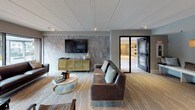 This community room is a thoughtfully designed, open-plan gathering space that seamlessly blends multiple functional zones into one cohesive environment. The lounge area serves as a focal point, anchored by a dramatic floor-to-ceiling stone tile accent wall on which a large flat-screen television is mounted. A gold-framed media console sits beneath the TV, complementing the rich brown leather sofas and accent chairs arranged around a marble-topped, gold-base coffee table atop a textured area rug.

Adjacent to the lounge, a substantial reclaimed wood dining table with upholstered chairs accommodates group gatherings or collaborative work sessions. A smaller bistro-style table with sculpted chairs is positioned near expansive floor-to-ceiling windows, offering views of the surrounding outdoor landscape and allowing generous natural light to fill the space.

A dedicated nook at the rear of the room features built-in upholstered bench seating on both sides of a central storage cabinet, accented by backlit wall niches in warm gold tones, providing an intimate retreat within the larger space. Track lighting throughout ensures the entire room is well-illuminated. The flooring transitions between stone-look tile and patterned carpet, tying the varied zones together with a modern, sophisticated aesthetic.