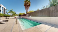 Located at 45 E 12th St in National City, California, this outdoor amenity area offers a well-maintained communal pool and surrounding recreation space designed for relaxed, open-air enjoyment. The pool itself features clear turquoise water, dual stainless steel entry railings, and tiled trim along its perimeter, providing both functionality and a clean aesthetic. The pool deck is finished in neutral concrete, offering ample space for lounge chairs and casual seating arrangements positioned to take full advantage of the abundant Southern California sunshine. A tall wooden privacy fence encloses one side of the space, creating a sense of seclusion, while mature palm trees and lush greenery add a tropical ambiance throughout. On the opposite side, a modern multi-story building with floor-to-ceiling glass windows, wood-accented facades, and private balconies serves as an impressive architectural backdrop. Shaded seating areas with umbrellas and outdoor dining furniture are situated nearby, making the space suitable for both lounging and socializing. Safety equipment, including a life ring, is visibly stationed poolside. The overall atmosphere is bright, open, and inviting, reflecting the relaxed lifestyle of the greater San Diego region. This thoughtfully designed outdoor space balances privacy, greenery, and modern architecture to deliver a comfortable and attractive recreational environment.