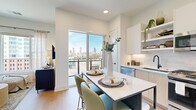 This thoughtfully laid-out one-bedroom, one-bathroom residence offers a efficient and modern living experience across a single floor. Light wood flooring flows throughout the main living area, anchored by a contemporary open-concept kitchen that serves as the heart of the home. The kitchen is outfitted with two-tone cabinetry—combining white upper cabinets with light wood lower cabinets—stainless steel appliances including a gas range, built-in microwave, and French door refrigerator, plus a subway tile backsplash and under-cabinet lighting. A freestanding island with a white quartz-style top and seating for two provides a casual dining space. A large sliding glass door opens to a private balcony, offering striking city skyline views and abundant natural light. The bedroom is a serene retreat, featuring a soft accent wall in muted sage green, plush carpeting, and a generous window with urban views. It connects directly to a dedicated walk-in closet fitted with custom built-in shelving, drawers, and hanging space. The bathroom is clean and modern, highlighted by a glass-enclosed walk-in shower with white subway tile surround, mosaic tile flooring, and matte black fixtures throughout.