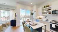 This thoughtfully laid-out one-bedroom, one-bathroom residence offers a efficient and modern living experience across a single floor. Light wood flooring flows throughout the main living area, anchored by a contemporary open-concept kitchen that serves as the heart of the home. The kitchen is outfitted with two-tone cabinetry—combining white upper cabinets with light wood lower cabinets—stainless steel appliances including a gas range, built-in microwave, and French door refrigerator, plus a subway tile backsplash and under-cabinet lighting. A freestanding island with a white quartz-style top and seating for two provides a casual dining space. A large sliding glass door opens to a private balcony, offering striking city skyline views and abundant natural light. The bedroom is a serene retreat, featuring a soft accent wall in muted sage green, plush carpeting, and a generous window with urban views. It connects directly to a dedicated walk-in closet fitted with custom built-in shelving, drawers, and hanging space. The bathroom is clean and modern, highlighted by a glass-enclosed walk-in shower with white subway tile surround, mosaic tile flooring, and matte black fixtures throughout.