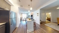 This single-floor residence presents a thoughtfully designed open-concept layout that seamlessly connects the kitchen, dining, and living areas. The kitchen is a standout feature, outfitted with dark wood cabinetry, stainless steel appliances including a refrigerator, range, microwave, and dishwasher, complemented by granite countertops and a white peninsula with bar seating for two. Pendant lighting and a rail track system illuminate the space with both style and function, while wood-look flooring runs throughout the main living areas.

The dining area accommodates an intimate round table and sits adjacent to the kitchen peninsula, creating a natural flow between spaces. The living room is characterized by soft blue-gray walls, a ceiling fan, and large sliding glass doors that flood the room with natural light and provide access to a balcony with exterior views.

The bedroom offers a calm, restful atmosphere with soft blue accent walls, a ceiling fan, and a window with blinds. The adjoining space includes a dresser and full-length mirror, providing practical storage and functionality. The overall design balances contemporary finishes with warm, neutral tones throughout.