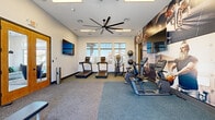 This fitness center is a thoughtfully designed single-floor facility that provides a comprehensive workout environment. The space is defined by its clean white walls, neutral carpet flooring, and an abundance of natural light streaming in through multiple windows, which offer views of the surrounding outdoor area. A large industrial-style ceiling fan anchors the room, ensuring proper air circulation throughout.

The cardio zone features multiple treadmills and an elliptical trainer lined along the windowed wall, complemented by stationary bikes and recumbent cycle machines. For strength training, the space is equipped with a cable machine, a weight bench, a dumbbell rack, and wall-mounted TRX suspension straps, providing a diverse range of exercise options. Stability balls are neatly organized on a wall-mounted rack, adding to the functional training offerings.

Large, vibrant fitness-themed photographic murals cover a prominent accent wall, creating an energizing and motivational atmosphere. Multiple flat-screen televisions are mounted on the walls for entertainment during workouts. Full-length mirrors line one wall, enhancing the sense of space and allowing users to monitor their form. A secondary door provides additional access and connects to an adjacent utility or restroom area. Overall, the facility delivers a modern, functional, and motivating environment for a full-body workout experience.