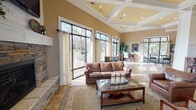 Park Lake's clubhouse is a single-floor gathering space designed for both relaxation and social events. The open-concept layout is anchored by a generously sized lounge area featuring soaring coffered and vaulted ceilings, rich tile flooring, and an abundance of natural light streaming through large black-framed windows and glass doors. A striking stacked-stone fireplace with a white mantle serves as a focal point in the lounge, complemented by leather seating, a glass-top coffee table, and warm neutral tones throughout.

Adjacent to the lounge, a dedicated dining area accommodates multiple round tables draped in linen, surrounded by mixed seating and set beneath an elegant coffered ceiling with recessed lighting. This space benefits from the same expansive windows that offer views of the surrounding landscape and direct access to a wraparound outdoor deck.

A built-in bar area with bar stools and decorative tile backsplash provides an ideal spot for entertaining. Throughout the space, consistent warm beige wall tones, tile flooring, and carefully placed decorative accents create a cohesive and inviting atmosphere. The outdoor deck, accessible from multiple points, extends the usable space and offers open views of the lake and surrounding natural scenery.
