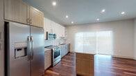 This single-floor residence showcases a contemporary design with a thoughtful layout and quality finishes throughout. The kitchen serves as the focal point of the living space, featuring stainless steel appliances including a French door refrigerator, electric range, built-in microwave, and dishwasher. White upper cabinetry contrasts with warm wood-toned lower cabinets, while a classic subway tile backsplash adds texture to the space. A granite-topped center island provides additional prep and storage space, complementing the matching granite countertops along the perimeter. Rich, dark hardwood flooring flows throughout the main living area, adding warmth and depth. A large sliding glass door floods the kitchen and living area with natural light and offers access to an outdoor space with exterior views. The bedroom is finished with plush carpeting and features a walk-in closet equipped with built-in shelving and hanging rods, providing generous storage. The bathroom is finished in neutral tones with large-format wall tiles surrounding a soaking tub and shower combination, chrome fixtures, and a granite-topped vanity, creating a clean and functional space.