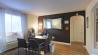 This single-floor space includes a well-appointed office area that combines functional design with distinctive stylistic choices. The room features a bold, dark chocolate-brown accent wall that serves as a striking focal point, complemented by lighter cream-toned surrounding walls that balance the overall aesthetic. A large, ornately framed rectangular mirror is mounted prominently on the accent wall, enhancing the sense of depth and reflecting natural light from the nearby window dressed with patterned curtains.

The workspace is centered around a dark wood desk accompanied by cushioned office chairs, providing a practical and professional setup. Warm hardwood flooring runs throughout, adding texture and a natural element to the space. A lighted vanity-style mirror stand positioned near the window offers additional ambient lighting alongside a tall floor lamp, ensuring the room is well-lit. An arched doorway on the far wall adds an architectural detail that softens the room's overall feel. Built-in shelving and organizational accessories visible near the desk contribute to the room's functionality. A dark area rug anchors the seating area, adding warmth and definition to the workspace.