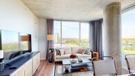 This contemporary single-floor residence showcases a striking blend of industrial and modern design elements throughout. The living room is a standout space, defined by expansive floor-to-ceiling windows that frame panoramic views of the surrounding cityscape and lush treetops, flooding the room with natural light. An exposed concrete ceiling and a signature structural concrete column add raw, architectural character, while warm hardwood-style flooring grounds the space with an inviting contrast.

The kitchen is well-appointed with a two-tone cabinet design — crisp white uppers paired with soft grey lowers — complemented by a classic white subway tile backsplash, white quartz countertops, and a full suite of stainless steel appliances including a range, over-the-range microwave, and refrigerator.

The bedroom continues the concrete ceiling aesthetic, with plush carpeting underfoot and large windows dressed in elegant drape panels, offering both comfort and style. Two bathrooms serve the residence: a full bath featuring grey cabinetry, a white countertop vanity, and a glass-enclosed shower-tub combination, and a second, more compact bath with a matching vanity and clean, minimal finishes. A well-proportioned hallway, adorned with a curated gallery wall of black-and-white framed artwork, connects the spaces with a refined, cohesive touch throughout.