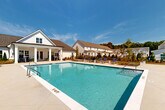 Located at 10 Bridgestone Ave in Conyers, Georgia, this community amenity space is centered around a generously sized resort-style swimming pool that serves as the focal point of an expansive outdoor retreat. The pool area features a graduated depth design and is surrounded by a wide, light-toned concrete deck outfitted with rows of blue-and-white lounge chairs and shaded umbrella stations, ideal for relaxing under the Georgia sun.

A standout feature of the space is the white-painted covered pavilion, designed in a classic farmhouse architectural style with peaked rooflines, exposed wood trusswork, and a ceiling fan for added comfort. The pavilion provides a shaded gathering area complete with outdoor seating, a mounted television, and views directly overlooking the pool.

Adjacent to the pool, the grounds open up to a well-maintained green lawn bordered by manicured landscaping beds, young trees, and ornamental plantings. A paved walkway winds through the landscaped area, connecting the various amenity zones within the community. Black iron fencing frames the perimeter, providing a clean boundary against the backdrop of the surrounding residential buildings. The entire space is thoughtfully designed to encourage outdoor living and community interaction.