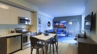 This single-floor space showcases a thoughtfully designed open-concept layout that seamlessly connects the kitchen, dining, and living areas into one cohesive and inviting environment. The kitchen is outfitted with stainless steel appliances, including a gas range with an overhead microwave, and features clean white cabinetry that maximizes storage while maintaining a minimalist aesthetic. Light hardwood flooring runs throughout, lending warmth and continuity to the space. The dining area comfortably accommodates a square table with modern curved chairs, positioned to allow easy flow between the kitchen and living room. The living room serves as a vibrant focal point, anchored by a bold blue sectional sofa, a round white coffee table, and a large statement artwork on the feature wall, which is painted in a contrasting grey tone. Ambient and accent lighting, including a large arc floor lamp and a contemporary wall-mounted lamp, enhance the dynamic atmosphere. The overall design balances a cool, modern color palette of blues, greys, and whites with warm natural wood elements, resulting in a stylish and functional living environment suited for both everyday living and entertaining.