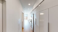 This single-floor unit showcases a clean, contemporary design characterized by an all-white interior palette, warm hardwood flooring, and recessed lighting throughout. The open-concept layout connects the living area to the kitchen, creating a seamless and functional flow.

The kitchen is thoughtfully designed with high-gloss white cabinetry extending to the ceiling, offering generous storage. Stainless steel appliances — including a range, built-in microwave, dishwasher, and refrigerator — lend a polished, modern feel. White quartz countertops and a tiled backsplash complete the space with crisp, cohesive detailing.

The living area is bright and open, filled with natural light streaming through large floor-to-ceiling windows that frame views of the surrounding urban neighborhood and tree canopy. The space offers a sense of openness and versatility.

The bathroom maintains the unit's minimalist aesthetic, featuring a white vanity with an undermount sink, white quartz countertop, a large mirror with overhead lighting, and a full tub and shower with clean white tile surround. Hardwood flooring carries through the space, reinforcing design continuity.

A corridor with additional built-in cabinetry connects the unit's spaces efficiently, with ample storage integrated into the architecture. Overall, the unit presents a bright, move-in-ready environment with modern finishes and an urban outlook.
