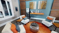 This single-floor space in Dallas presents a well-appointed lounge area designed with a contemporary aesthetic. The interior is characterized by a striking teal-blue accent wall that serves as a bold focal point, complemented by warm white and gray surrounding walls. Decorative wall panels in aged copper tones and textured artwork in gold and neutral hues add visual depth and dimension to the space.

The seating arrangement is thoughtfully composed, featuring white upholstered armchairs accented with orange and blue throw pillows, alongside an orange sofa that reinforces the room's vibrant color palette. A metallic hammered-finish ottoman, topped with a tic-tac-toe game board, anchors the seating area as both a functional and decorative centerpiece. A dark wood side table adds warmth and contrast.

An orange floating shelf mounted against the teal wall displays beverage accessories and glassware, with a water/beverage dispensing station positioned nearby, suggesting a hospitality-oriented function. A digital display panel is mounted near the entrance. The space features patterned area rugs layered over solid flooring, contributing to a dynamic, layered interior design.