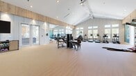 Located in Port Wentworth, Georgia, this fitness center offers a thoughtfully designed, single-floor layout that accommodates a full range of workout activities. The main gym area is characterized by soaring vaulted ceilings with white-painted shiplap paneling and warm wood accent banding, creating an open and airy atmosphere. Large, multi-pane windows line the walls, flooding the space with natural light and providing views of the surrounding landscape. The main floor features durable wood-tone flooring and houses a comprehensive selection of equipment, including strength machines, cable systems, free weights, kettlebells, battle ropes, and a full row of cardio machines. A striking abstract wall mural in soft teal and blue tones serves as a standout design focal point. Floor-to-ceiling mirrors along one wall visually expand the space while supporting functional training. A dedicated studio or yoga room, finished with soft teal walls, white wainscoting trim details, and smooth flooring, provides a separate area for group fitness or stretching, featuring a wall-mounted TV, large mirrors, and ample space for balance and flexibility equipment. Ceiling fans throughout promote air circulation, and recessed lighting supplements the natural light for consistent illumination across all areas.