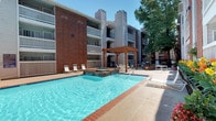 This outdoor pool area serves as a central amenity, thoughtfully designed for both relaxation and recreation. The centerpiece is a generously sized swimming pool with clear, blue water framed by brick-trimmed edges and a smooth concrete pool deck. Adjacent to the pool sits an elevated spa, also finished with classic brick detailing, offering a distinct and inviting retreat within the same space.

A substantial wooden pergola anchors one side of the deck, casting a pattern of filtered shade across the seating area beneath. Outfitted with round patio tables and cushioned lounge chairs, this covered zone provides a comfortable space for gathering or unwinding outdoors. Additional chaise lounges are positioned around the pool deck to take full advantage of open sunlight.

The courtyard setting is enclosed by multi-story residential buildings featuring brick and siding exteriors, with private balconies overlooking the pool area. Mature trees rise above the roofline in the background, contributing to a sense of privacy and greenery. Raised planters filled with colorful flowering plants and well-maintained shrubs line the perimeter, enhancing the landscaped ambiance. A gated entry and posted pool rules signage complete the well-organized and welcoming outdoor environment.