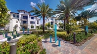 This compact outdoor fitness station is situated within a beautifully landscaped courtyard that serves as a communal amenity space. The area centers on a set of teal-colored outdoor exercise machines mounted on a herringbone-patterned brick paver surface, providing a dedicated spot for residents to engage in low-impact physical activity. The fitness equipment is compact yet functional, designed to accommodate stretching and resistance-based exercises.

Surrounding the station, the landscape is lush and tropical in character, featuring tall, mature palm trees that provide natural shade and a resort-like atmosphere. Neatly trimmed shrubs, flowering plants, and manicured green lawns border the paved area, contributing to the overall sense of a well-maintained environment. Bollard-style pathway lighting is positioned throughout the courtyard, ensuring usability during evening hours.

The backdrop consists of multi-story, Mediterranean-inspired residential buildings characterized by white stucco exteriors, arched architectural details, and red tile roofing accents. The open-air setting benefits from abundant natural sunlight and a clear sky, creating a bright and inviting outdoor atmosphere. The combination of thoughtful landscaping, vibrant equipment, and elegant surrounding architecture makes this fitness station an appealing and functional community feature.