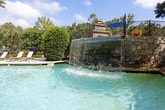 This inviting outdoor space centers around a well-maintained swimming pool, offering a tranquil retreat set within a beautifully landscaped environment. The pool area is framed by a substantial stacked-stone retaining wall, constructed from irregular-cut granite blocks that lend a timeless, classic character to the space. A sleek wrought iron fence tops the wall, providing both security and an open view of the surrounding greenery.

A generous pool deck runs alongside the water, lined with a row of sling-style lounge chairs ideal for relaxing in the sun. The pool itself features a curved, freeform shape and clear blue water, with a water feature visible at one end that adds both visual interest and ambient sound. Stone steps lead from the upper grounds down to the pool level, integrating the space naturally into the sloped terrain.

Mature trees provide generous shade and a sense of seclusion, while neatly trimmed shrubs, flowering plants, and ground cover soften the stone and concrete elements throughout. A multi-story building with classic architectural detailing is visible in the background, adding to the established character of the property. The overall ambiance is peaceful and resort-like, offering a well-appointed outdoor escape surrounded by natural beauty.