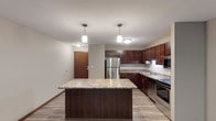 This single-floor residence centers around a thoughtfully designed kitchen that serves as the heart of the space. The kitchen features rich, dark-stained wood cabinetry extending along two walls, providing generous storage and a warm, grounded aesthetic. Granite-patterned countertops run throughout, complementing the deep tones of the cabinetry. A central island with a matching countertop surface offers additional workspace and casual dining potential, anchored beneath two cylindrical pendant lights that provide focused task lighting. Stainless steel appliances, including a French-door refrigerator, a built-in microwave, and a range with oven, deliver a modern, cohesive look. The kitchen sink is positioned along the perimeter counters, and under-cabinet lighting adds both practicality and ambiance. Light wood-toned flooring runs throughout, brightening the space and contrasting cleanly with the darker cabinetry. A textured ceiling and neutral wall tones contribute to a clean, understated interior. The entry door, finished in warm wood tones, is visible from the kitchen, reinforcing the open and connected layout of this single-level residence.