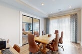 This single-floor meeting space is thoughtfully designed to accommodate professional gatherings in a comfortable and polished environment. At the heart of the room sits a substantial solid wood pedestal conference table, surrounded by brown leather executive chairs on casters, offering both style and practicality for collaborative work sessions.

Natural light flows generously into the space through large windows dressed with layered sheer curtains and patterned accent drapes, creating a bright yet comfortable atmosphere. A set of full-height glass sliding doors along one wall provides a seamless visual connection to an adjacent lounge or common area, enhancing the sense of openness while maintaining acoustic separation.

A wall-mounted flat-screen display serves as the room's primary presentation focal point, complemented by decorative framed artwork that adds warmth to the neutral-toned walls. Recessed ceiling lighting supplements natural light, ensuring consistent illumination throughout. A secondary workstation with dual monitors is positioned along one wall, offering additional functionality for hybrid or tech-driven meetings.

Warm-toned hardwood flooring runs throughout, tying together the room's inviting aesthetic. Crown molding and a tray ceiling detail lend a refined architectural character to the space. The overall design balances professional utility with an approachable, well-curated ambiance suited for meetings, presentations, or collaborative work.
